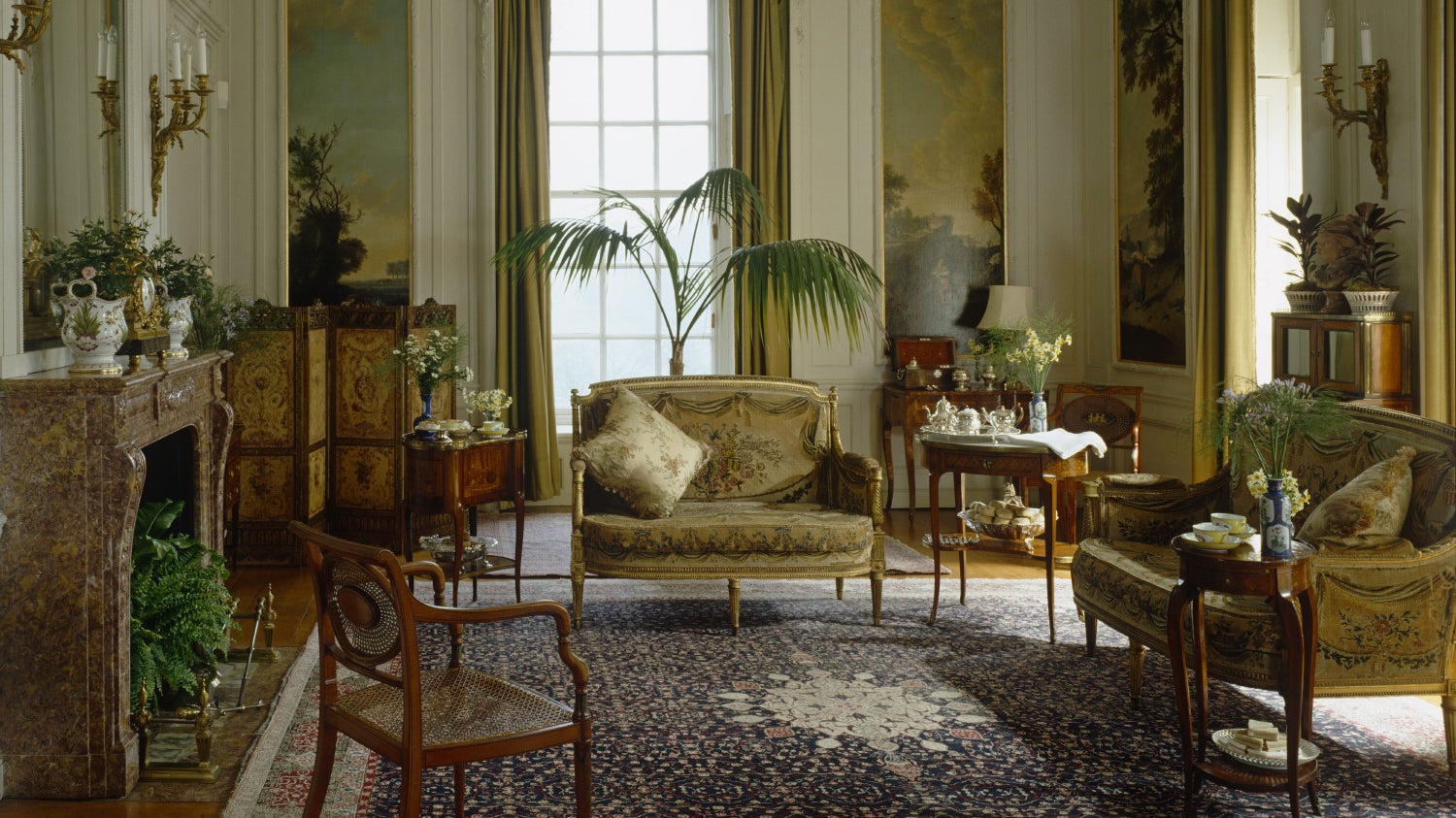 Elegant period drawing room with gold damask furniture, fireplace, ornate mirrors, potted plants, and tall windows with golden drapes