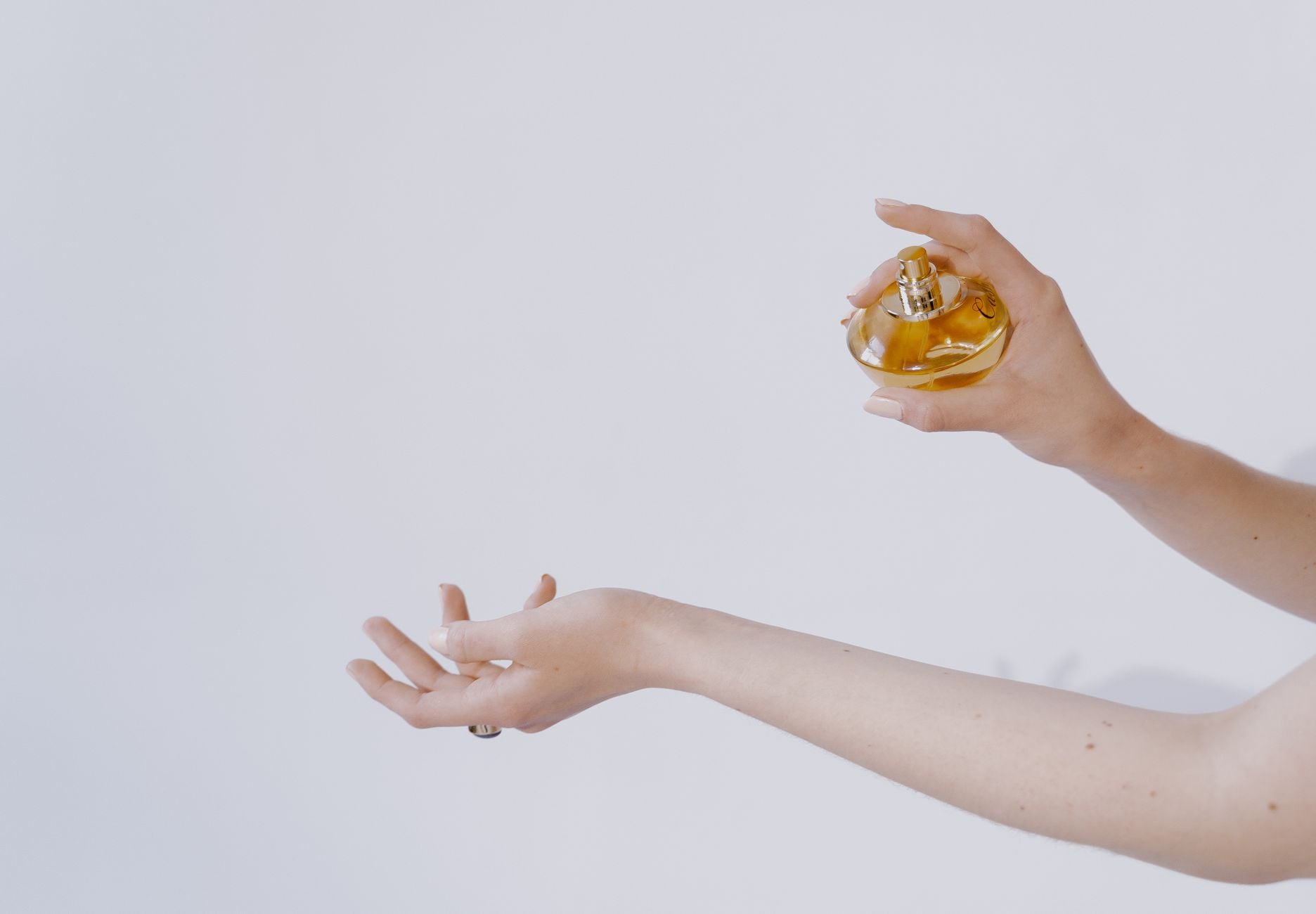 Kobieta trzymająca złotą butelkę perfum nad dłonią, minimalistyczne ujęcie na jasnym tle