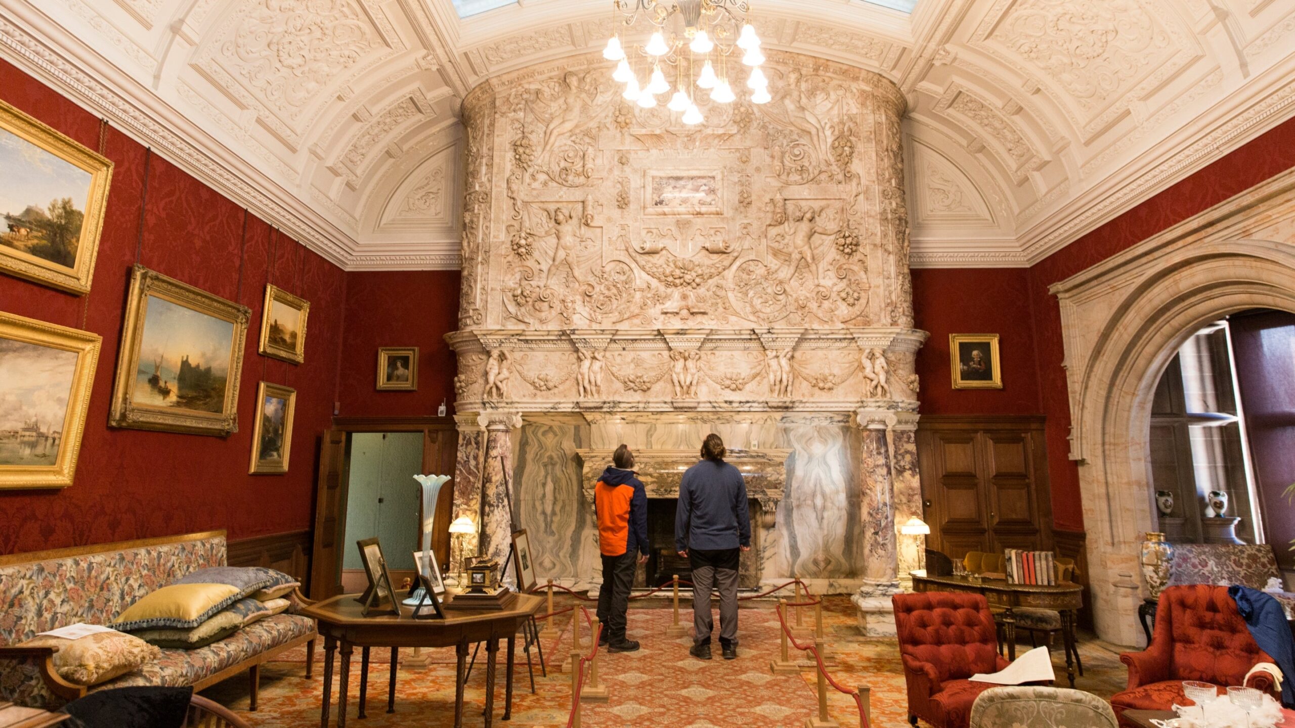 Grand fireplace with ornate carved marble surround in red-walled room with gold-framed paintings and period furniture