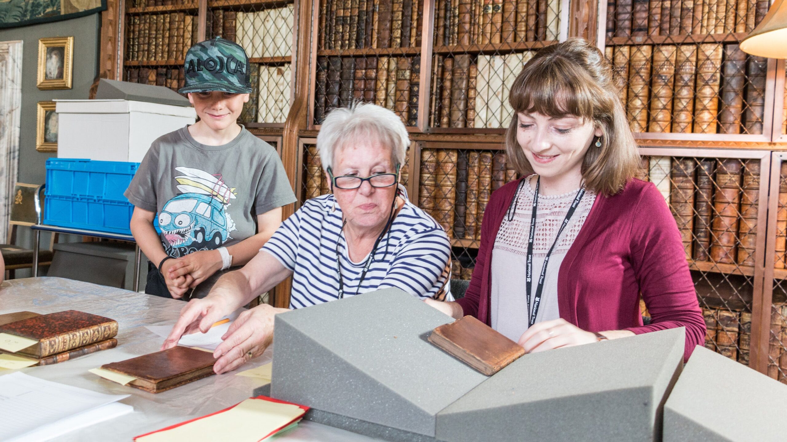 Woman and children examining antique books at a table in a library with wooden bookcases and leather-bound volumes