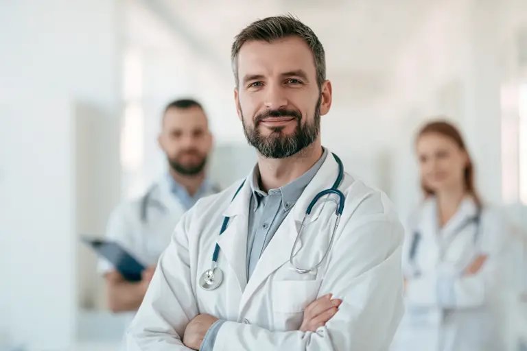 Lekarz w białym kitlu stoi z założonymi rękami, w tle dwaj inni pracownicy medyczni