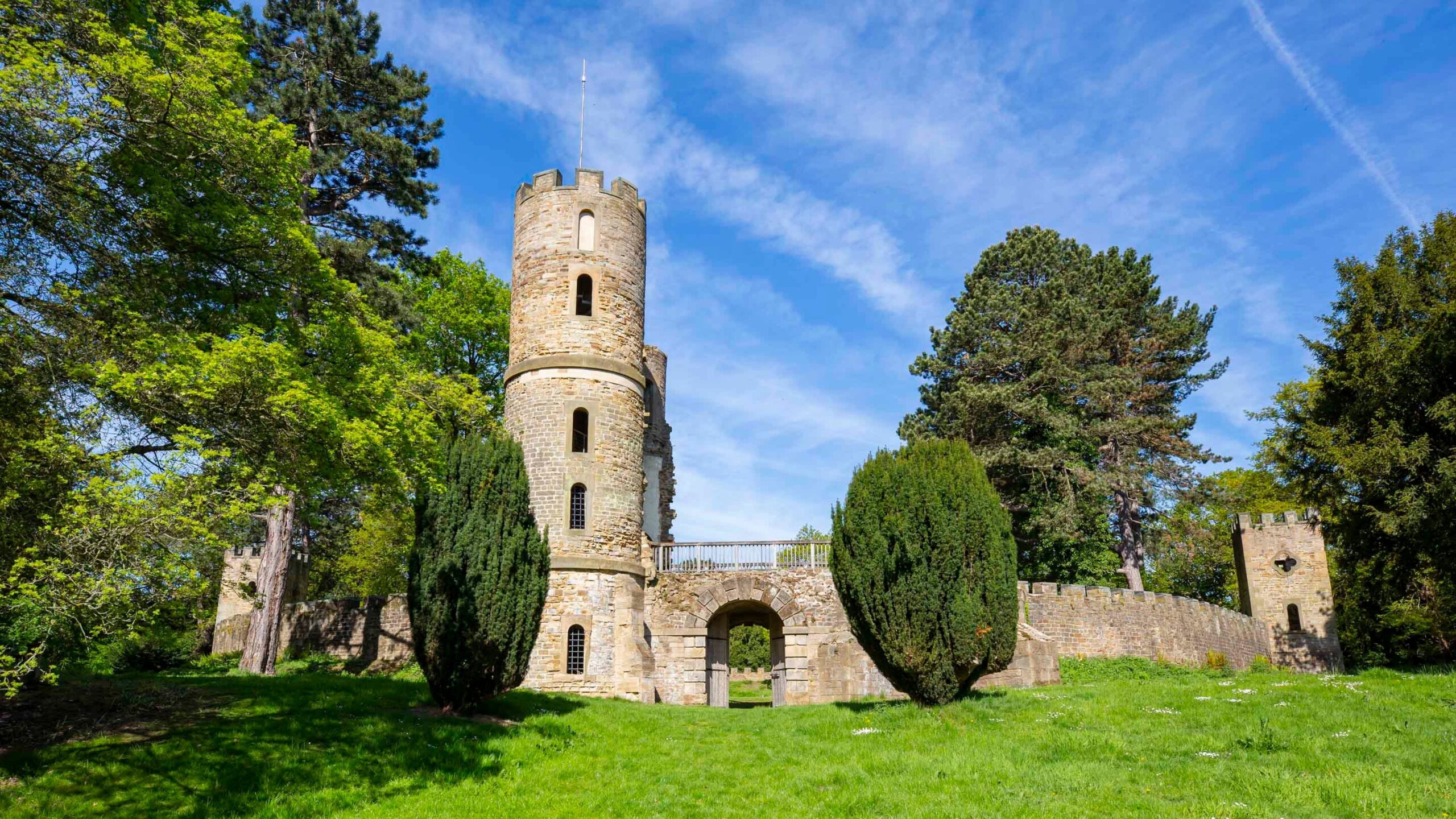 Stone castle tower with crenellations surrounded by cypress trees and green grass under blue sky