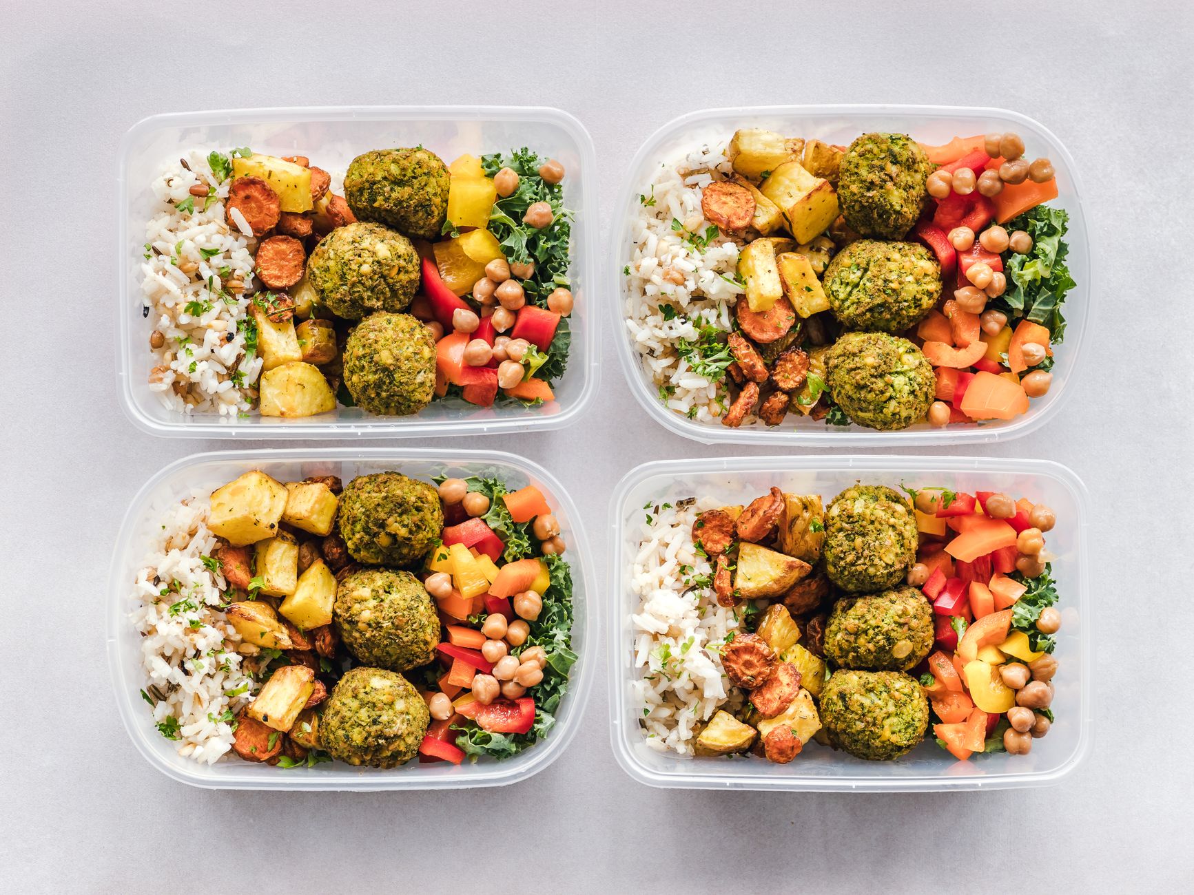 Cztery przezroczyste pojemniki z porcjami lunch boxów zawierające ryz, falafele, ziemniaki, kielbaskę, cieciorę, paprykę i sałatę