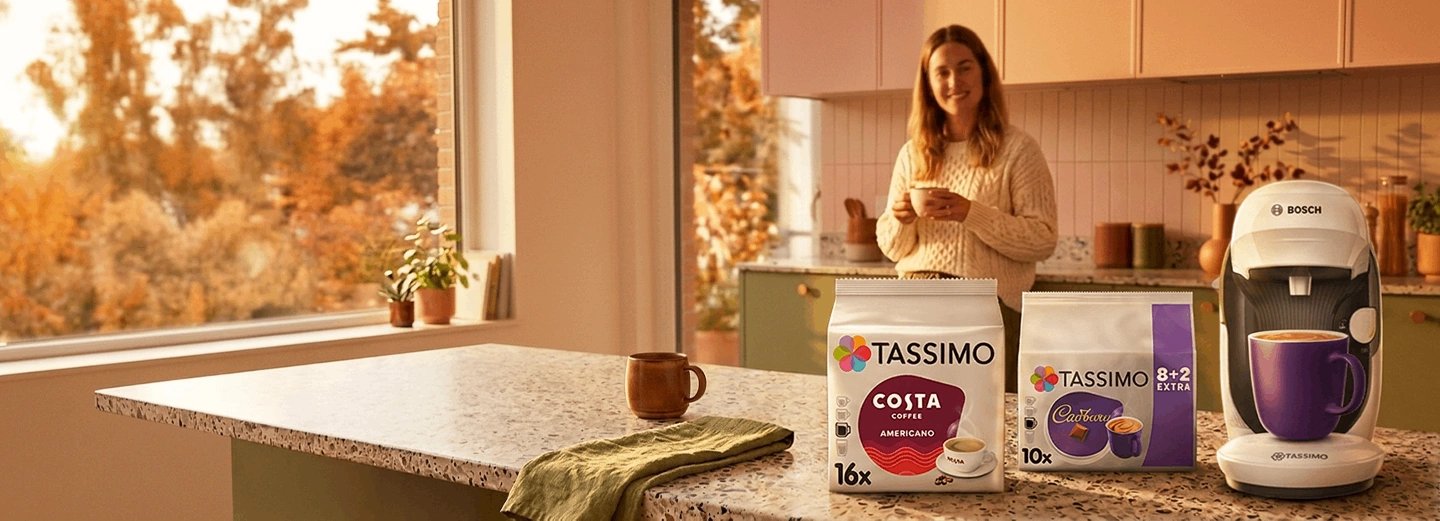 Woman in kitchen holding TASSIMO pod next to white Bosch machine, Costa and Cadbury T DISC boxes on counter with autumn window view